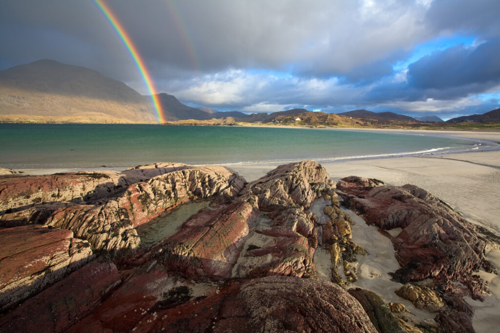 Rocky shoreline with red and gray rocks, sandy beach, and a double rainbow over a mountainous landscape under a partly cloudy sky.