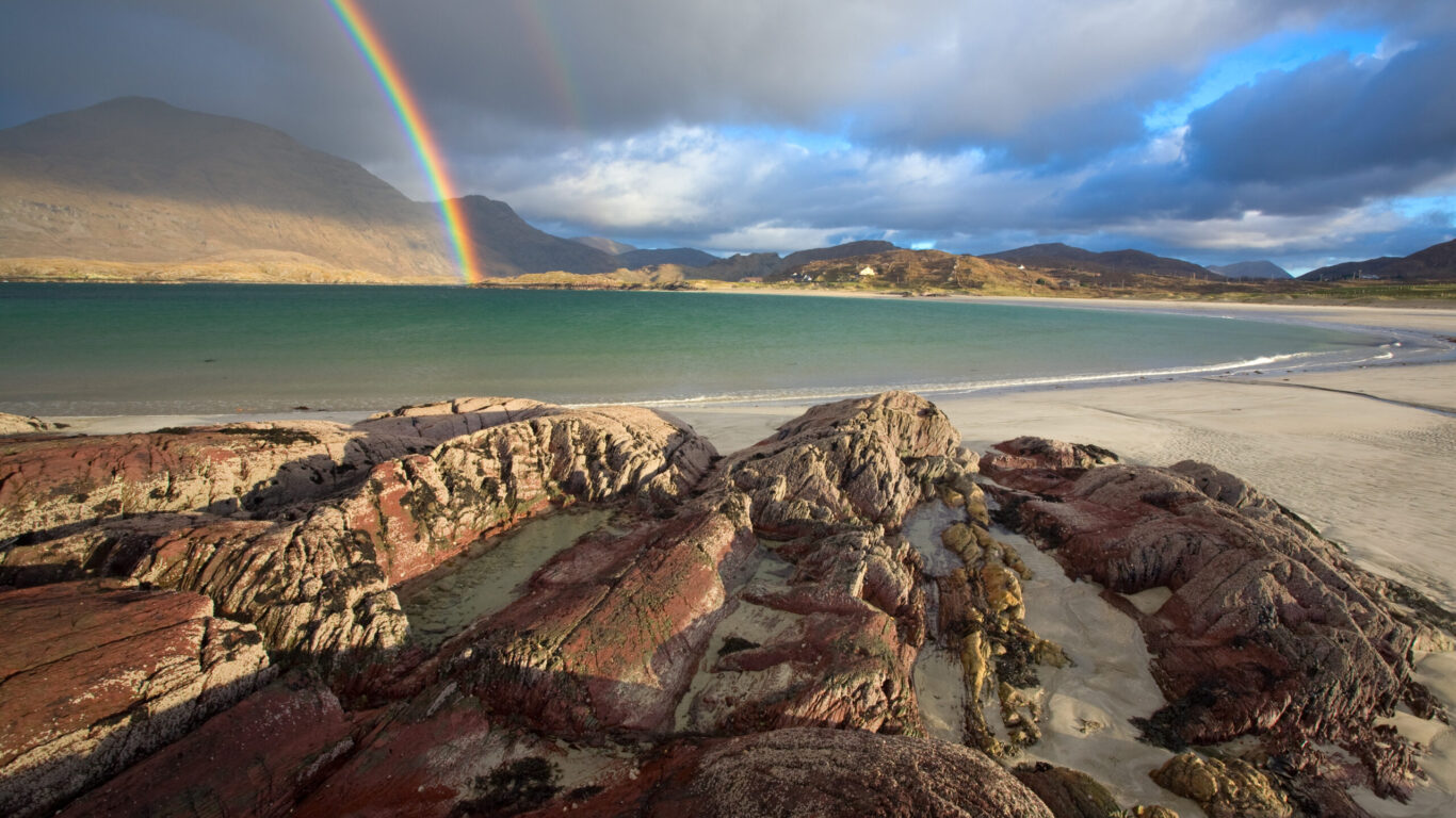 Rocky shoreline with red and gray rocks, sandy beach, and a double rainbow over a mountainous landscape under a partly cloudy sky.