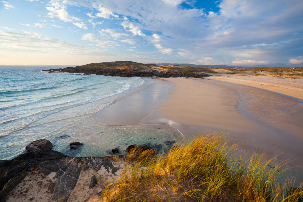 A sandy beach with gentle waves and scattered rocks, bordered by grassy dunes under a partly cloudy sky.