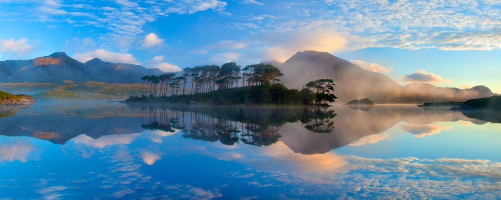 A calm lake reflects an island of trees, distant mountains, and a partly cloudy blue sky at sunrise or sunset. Mist hovers near the shoreline.