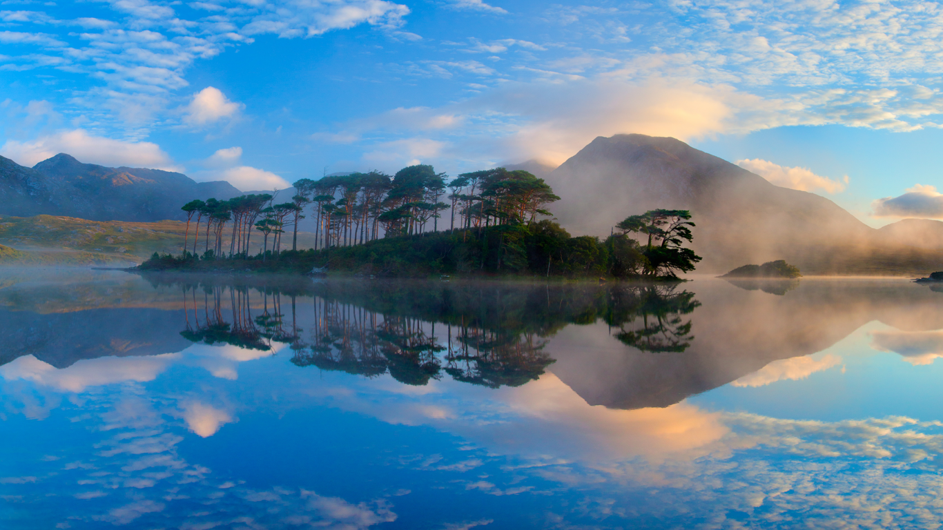 A calm lake reflects an island of trees, distant mountains, and a partly cloudy blue sky at sunrise or sunset. Mist hovers near the shoreline.