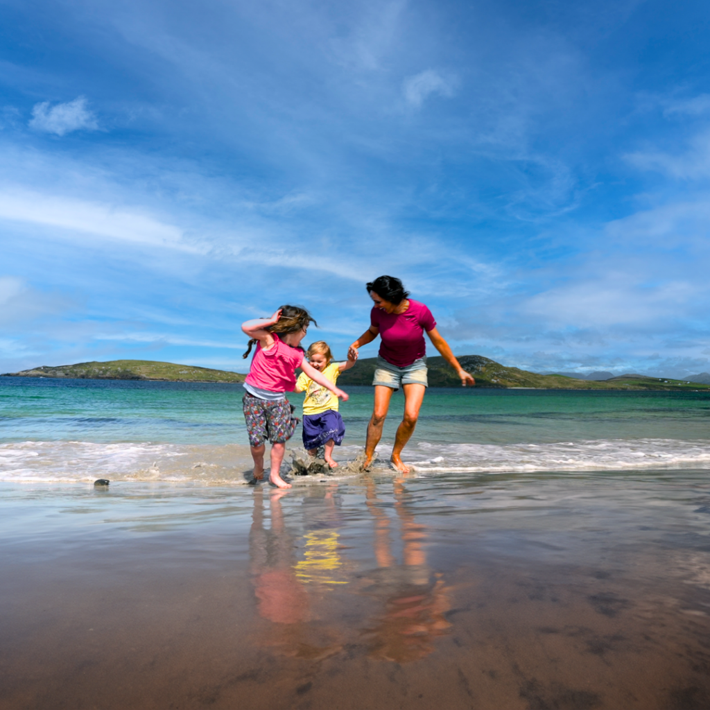 An adult and two children play and walk at the edge of the sea on a sandy beach with hills and blue sky in the background.