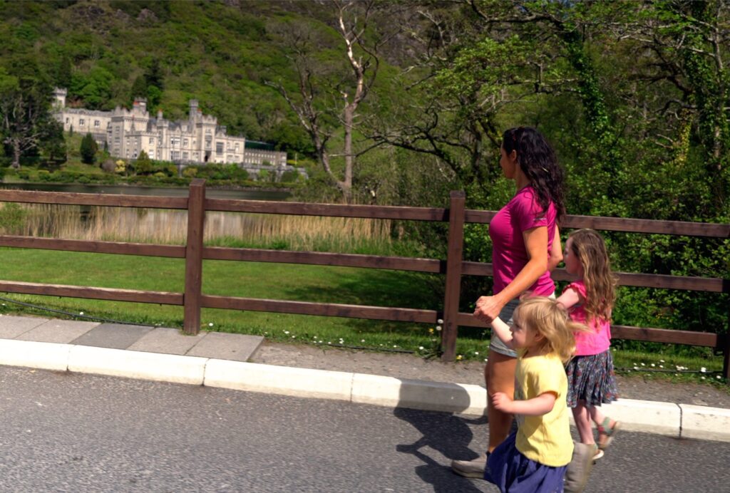 An adult and two children walk along a road by a wooden fence with a large stone building and trees visible across a lake in the background.