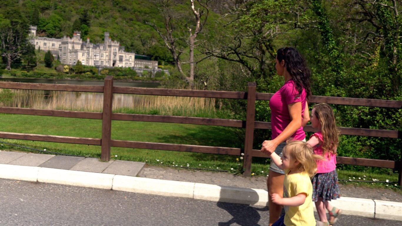 An adult and two children walk along a road by a wooden fence with a large stone building and trees visible across a lake in the background.