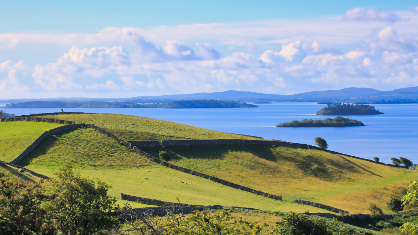 Rolling green hills bordered by stone walls overlook a calm blue lake with small islands under a partly cloudy sky.