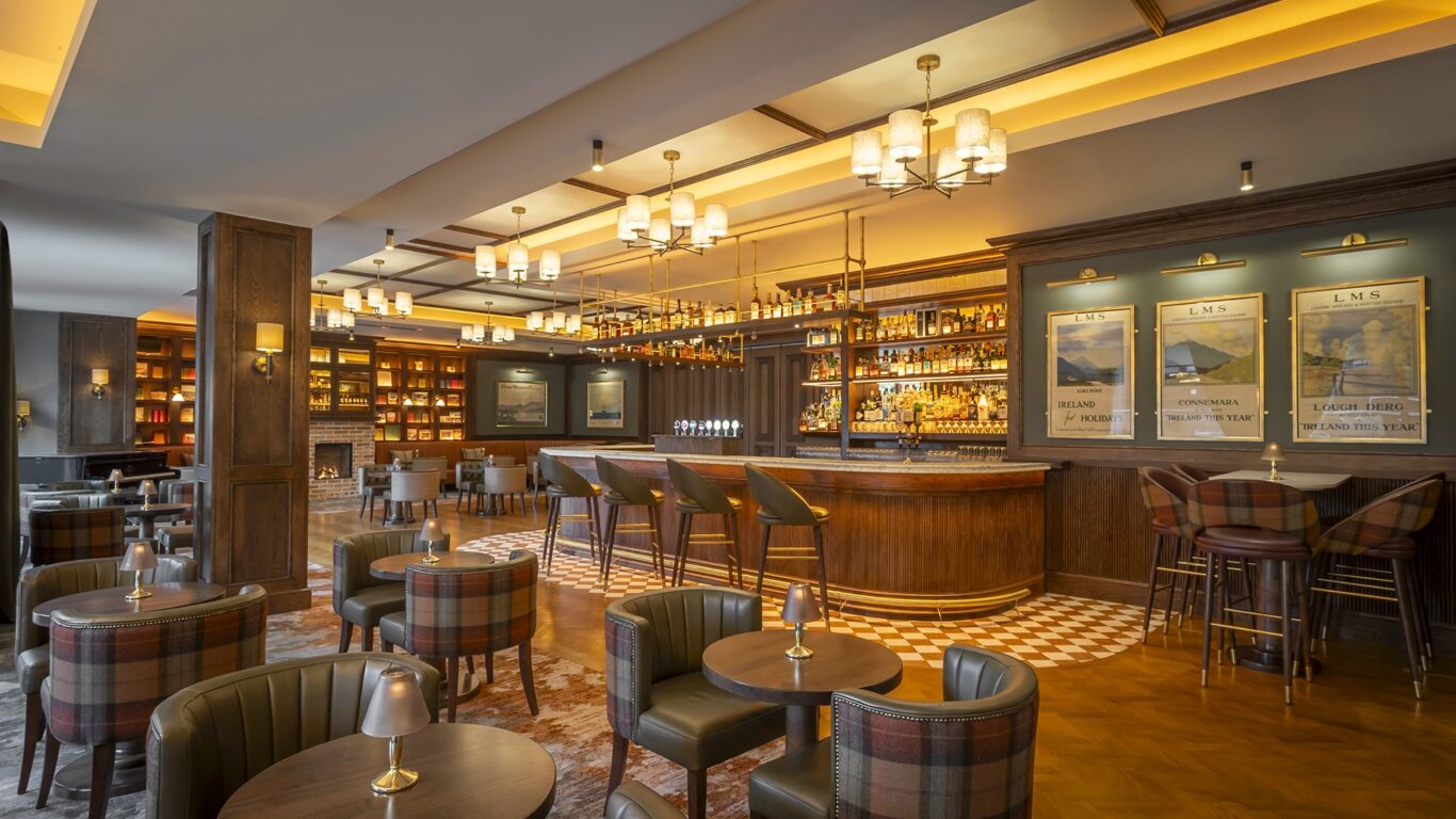 Upscale bar with wooden furnishings, round tables, upholstered chairs, and a central bar area with shelves of bottles behind it, warmly lit by ceiling and wall lights.