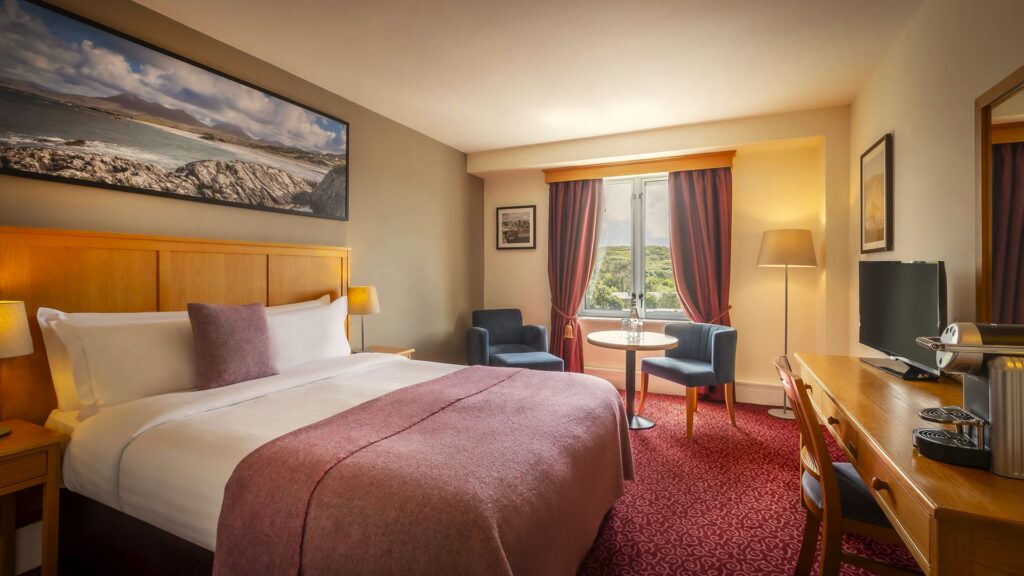 A hotel room with a double bed, two chairs and a round table by the window, a desk with a TV and coffee maker, and wall art above the bed.