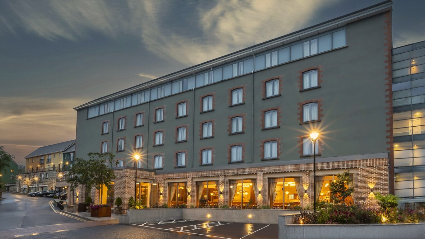 A modern four-story hotel building with large windows, illuminated warmly from inside at dusk, with adjacent parking spaces and landscaped entryway.