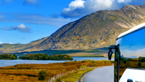 A white bus travels on a winding road beside a lake, with grassy fields and a mountain under a partly cloudy blue sky in the background.