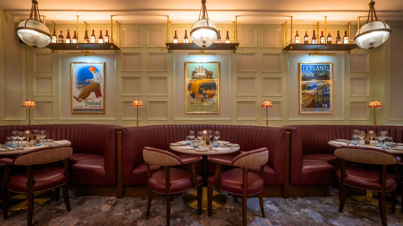A restaurant interior with red leather booths, round tables set for dining, wall lamps, vintage travel posters, and bottles displayed on a shelf above.