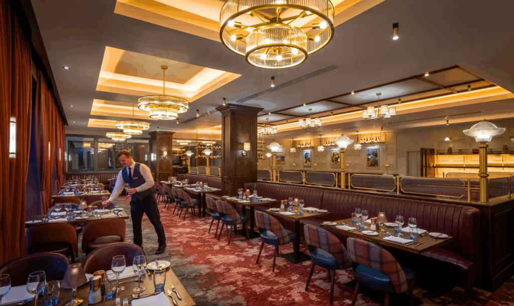 A waiter sets a table in a modern restaurant with brown leather booths, elegant lighting fixtures, and a fully set dining area.