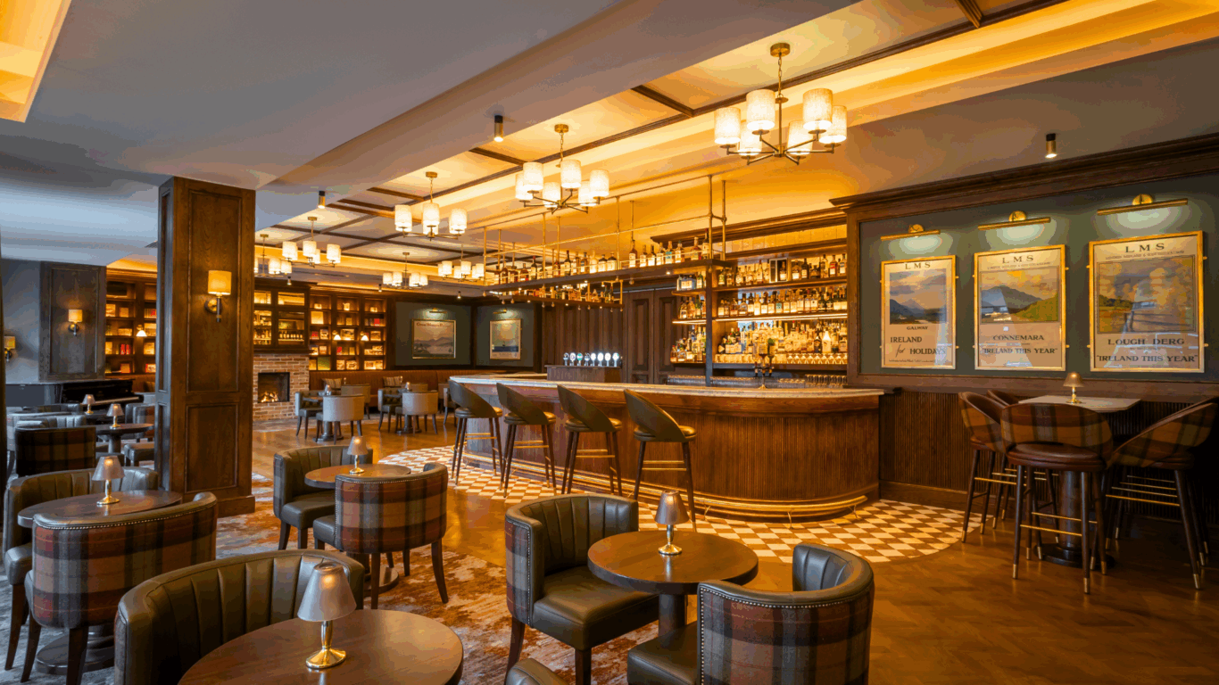 A stylish, modern bar with wooden decor, several round tables, plaid armchairs, and a well-stocked bar counter under warm lighting.