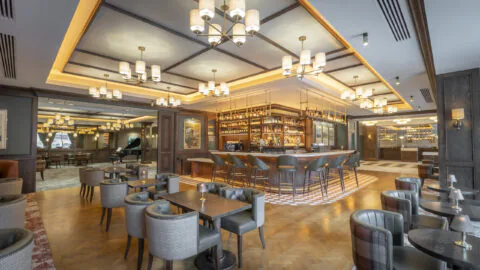 Modern bar and lounge area with wooden floors, central bar, upholstered chairs, small tables, and warm ceiling lighting.