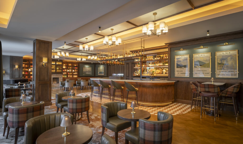 A modern, elegant bar with cushioned chairs, round tables, warm lighting, and shelves stocked with bottles behind the bar counter.