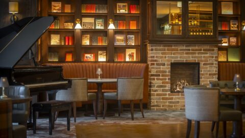 A cozy lounge with a grand piano, upholstered seating, a brick fireplace, and bookshelves filled with books and decorative items along the wall.