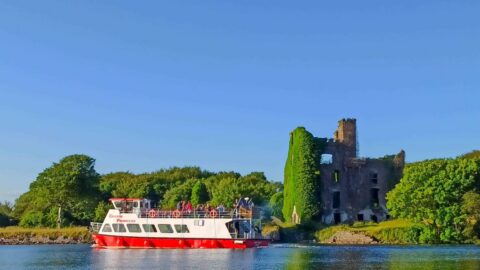 A red and white tour boat sails on a calm river near an ivy-covered stone ruin surrounded by green trees under a clear blue sky.