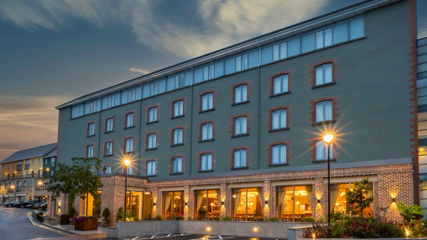 Four-story modern hotel with large ground-floor windows illuminated at dusk, surrounded by streetlights and a parking area.
