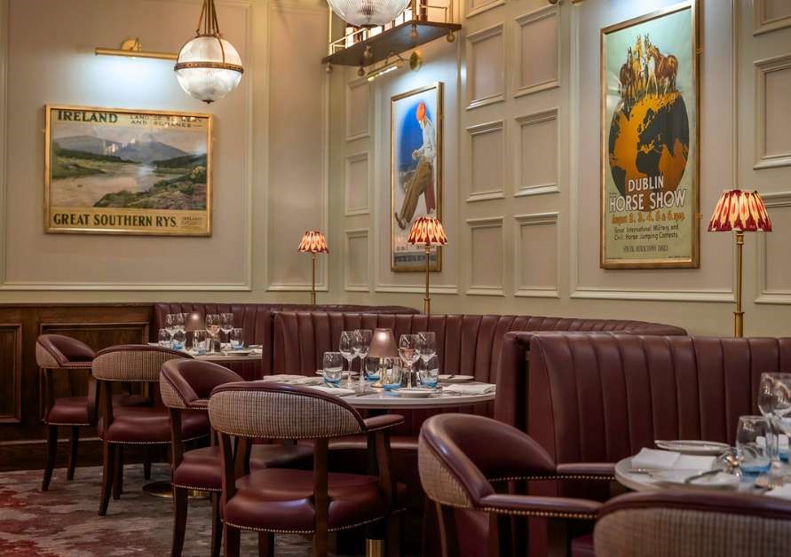 Restaurant interior with burgundy leather seating, set tables, vintage-style lamps, and framed travel posters on paneled walls.