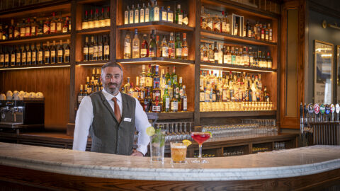 A bartender stands behind a curved marble bar with three drinks in front; shelves behind him hold a variety of liquor bottles and glasses.