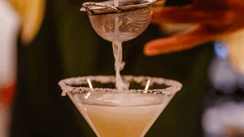A person strains a light-colored cocktail into a sugar-rimmed martini glass using a metal strainer.