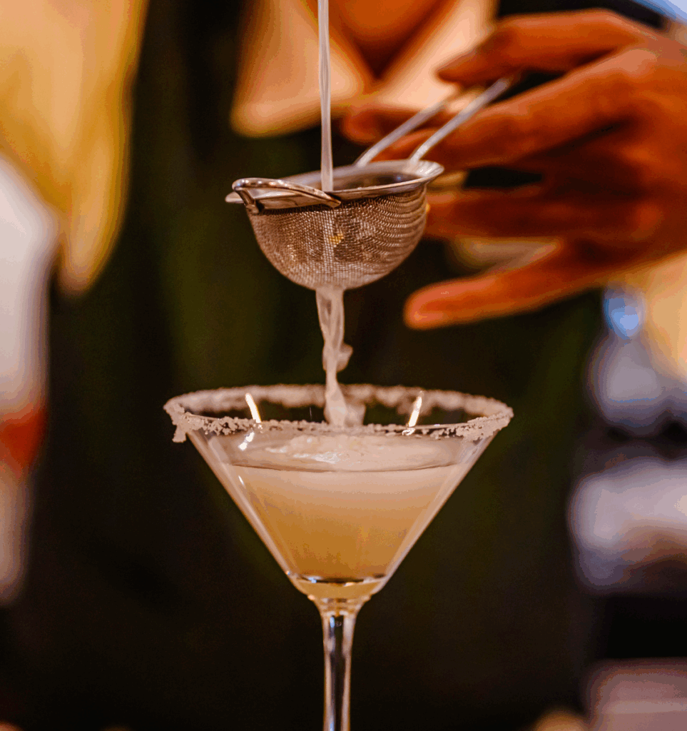 A person strains a light-colored cocktail into a sugar-rimmed martini glass using a metal strainer.