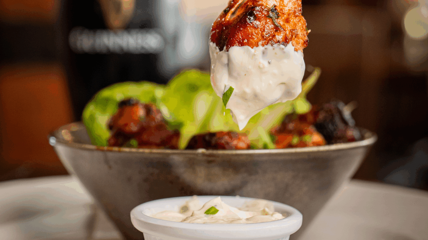 A hand dips a saucy chicken wing into a ramekin of white dipping sauce, with a bowl of more wings and a pint of Guinness in the background.