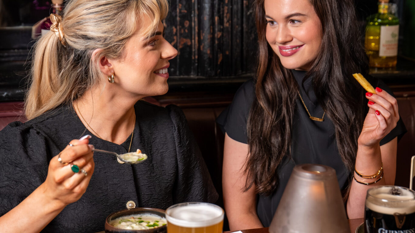 Two women sit at a restaurant table, eating and smiling at each other, with food, a pint of beer, and a glass of Guinness on the table.