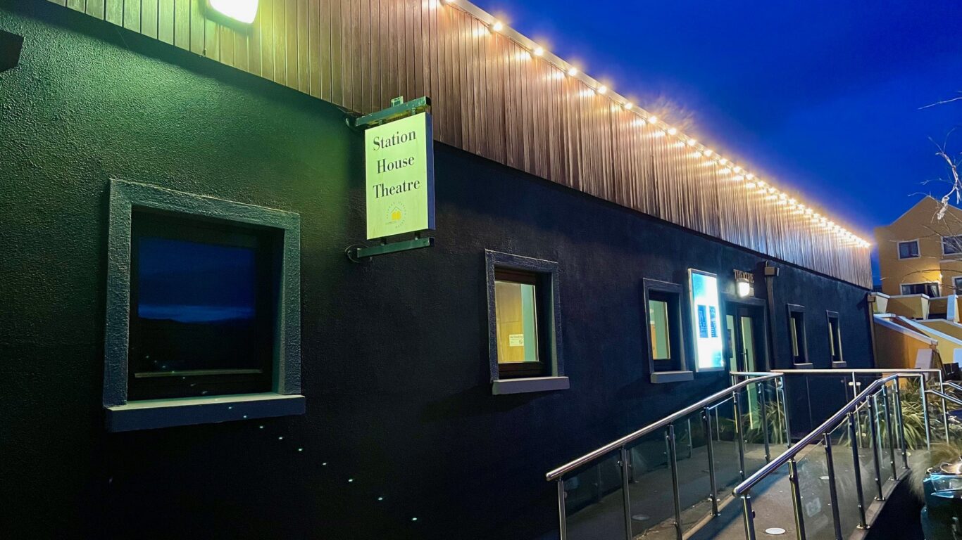 Exterior of the Station House Theatre at dusk, with illuminated sign, string lights, ramp with railing, and several windows visible.
