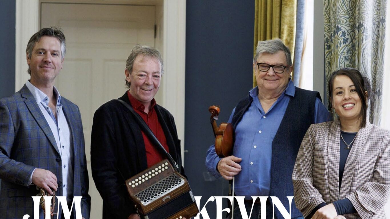Four adults stand indoors, posing for a photo. Two hold musical instruments: an accordion and a violin. They are smiling and dressed in semi-formal attire.