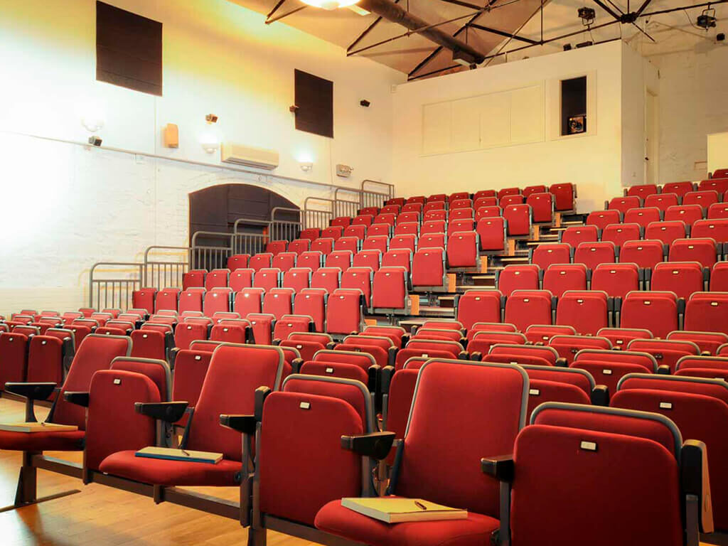 Rows of empty red theater-style seats in a small lecture hall with wooden floors, white walls, and metal railings.