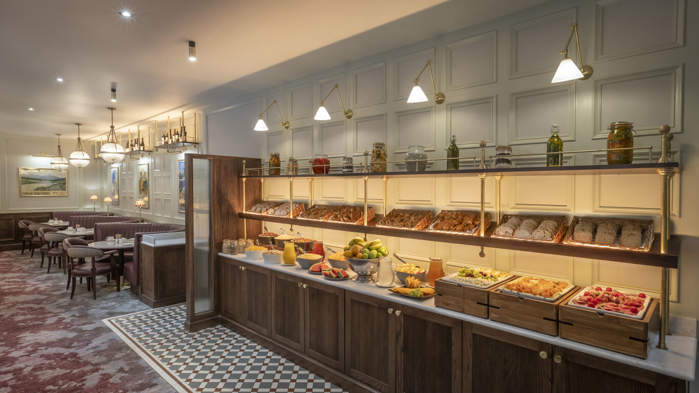 Buffet counter with assorted breads, fruits, and preserved items in a well-lit dining area with tables and upholstered chairs.