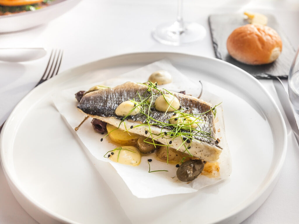 A cooked fish fillet topped with herbs and sauce, served on parchment paper with olives and vegetables, on a white plate next to a bread roll.