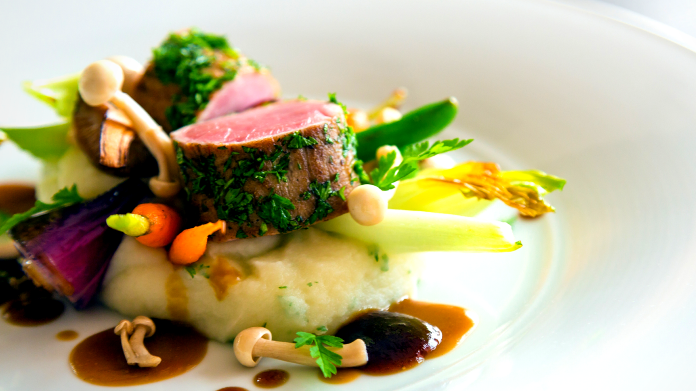 A plated gourmet dish featuring herb-crusted meat slices, mashed potatoes, assorted vegetables, mushrooms, and brown sauce on a white plate.