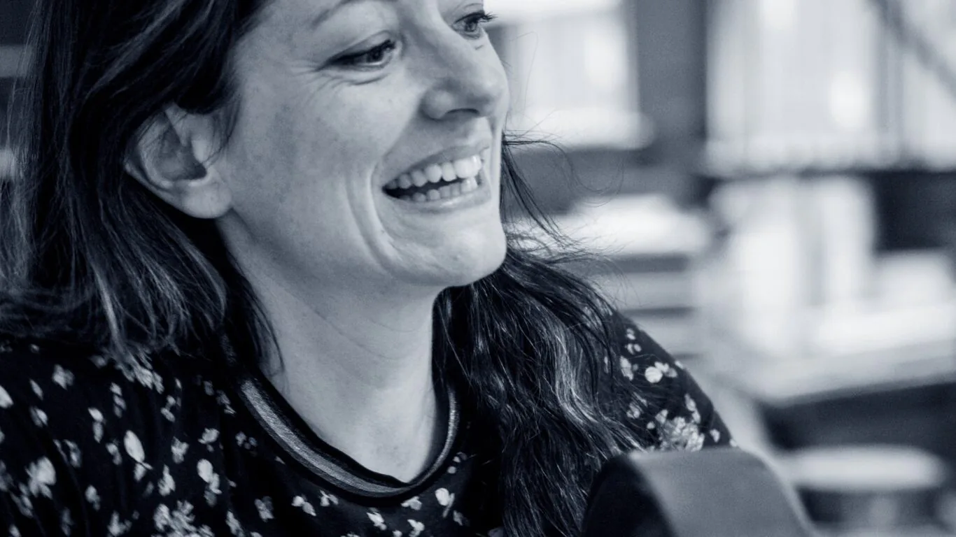 Woman with long hair and a floral shirt smiles while sitting indoors, with blurred shelves and objects in the background.