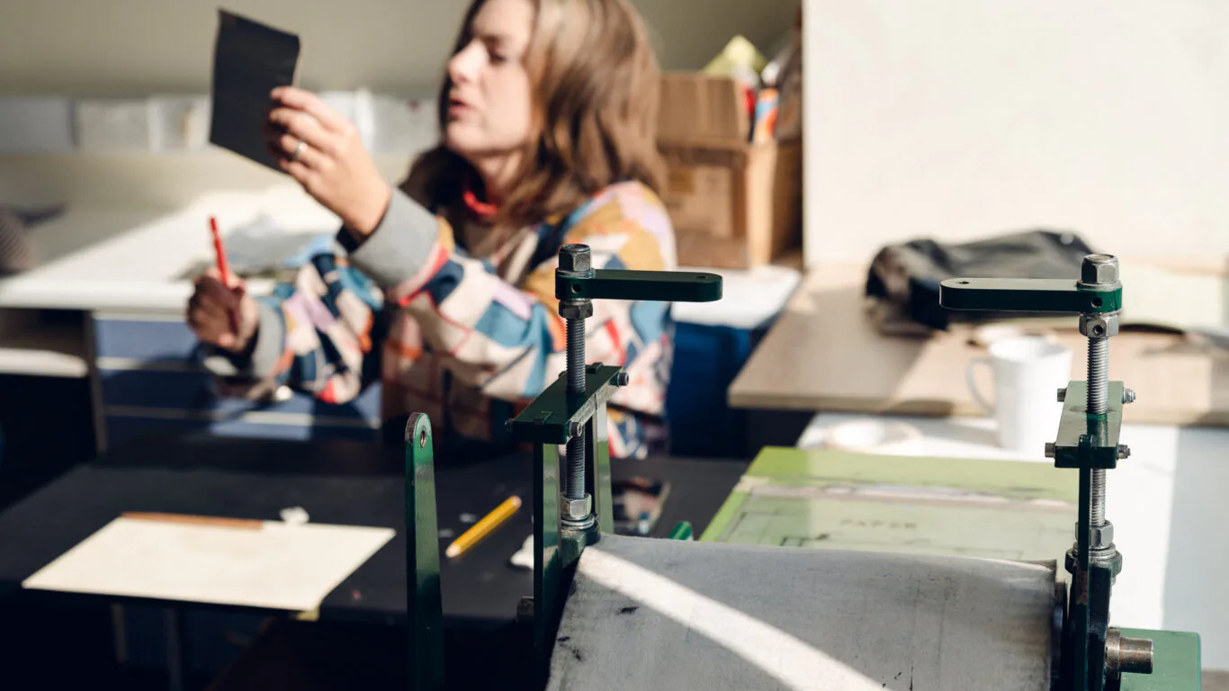 A person sits at a desk examining a black card, with art supplies and a printmaking press visible in the foreground.