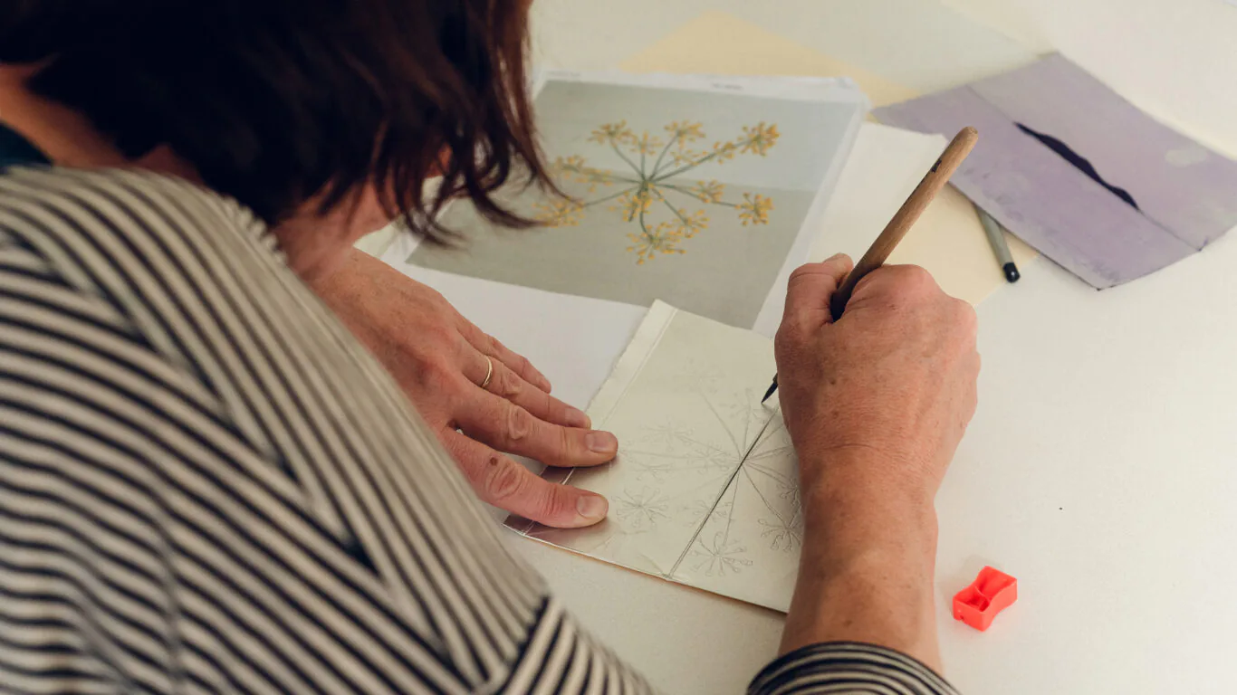 Person sketching a botanical design with a pencil on paper, referencing a printed illustration. Other drawing materials and templates are visible on the desk.