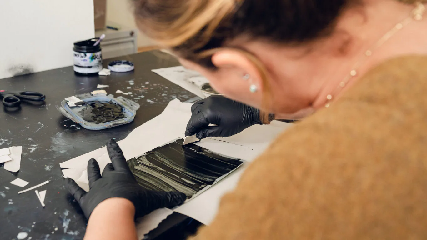 A person wearing black gloves uses a tool to spread black paint or ink on a rectangular surface over paper on a table.