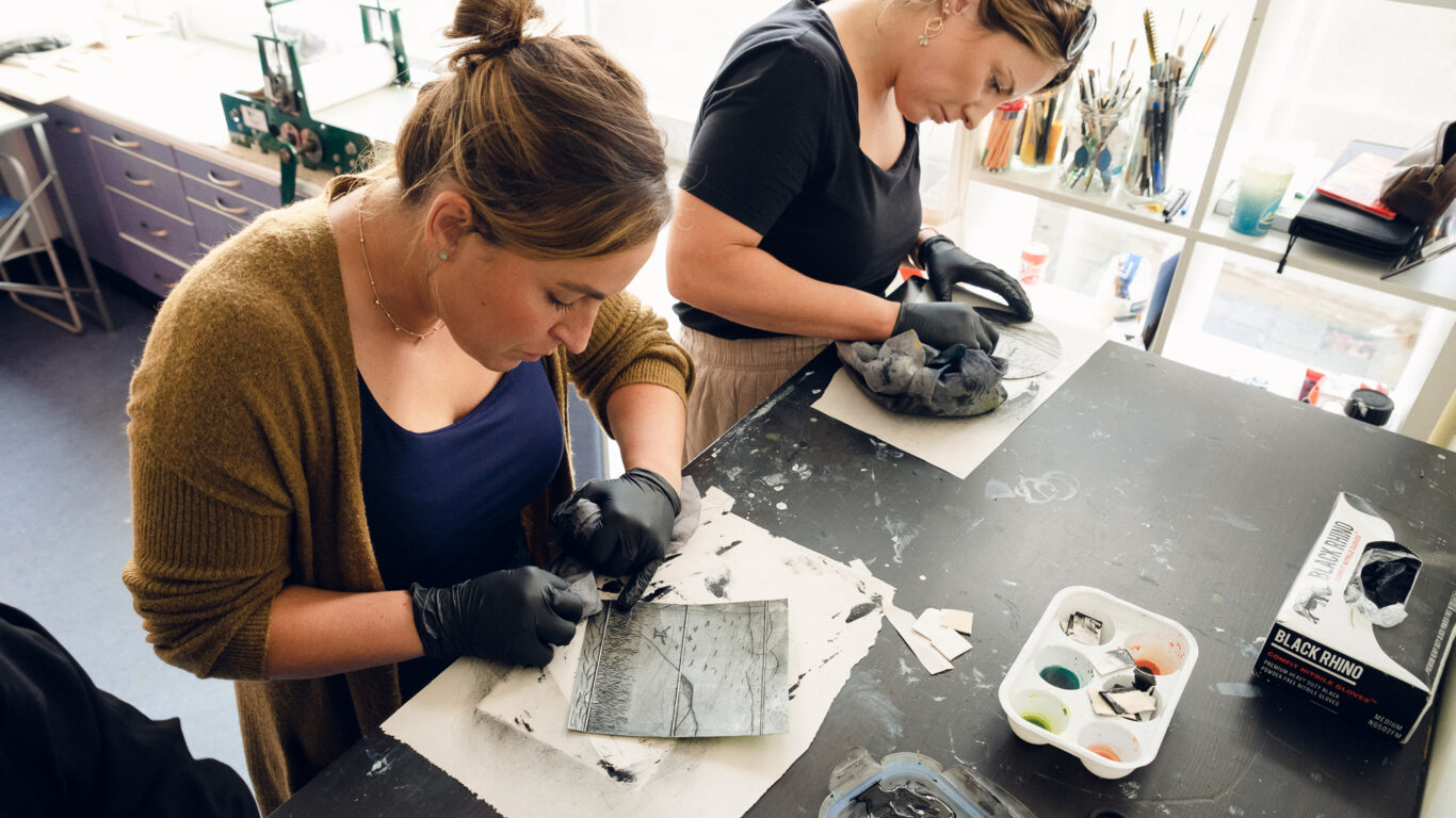 Two women wearing black gloves work intently on art projects at a table with ink, brushes, and various supplies in a well-lit studio.