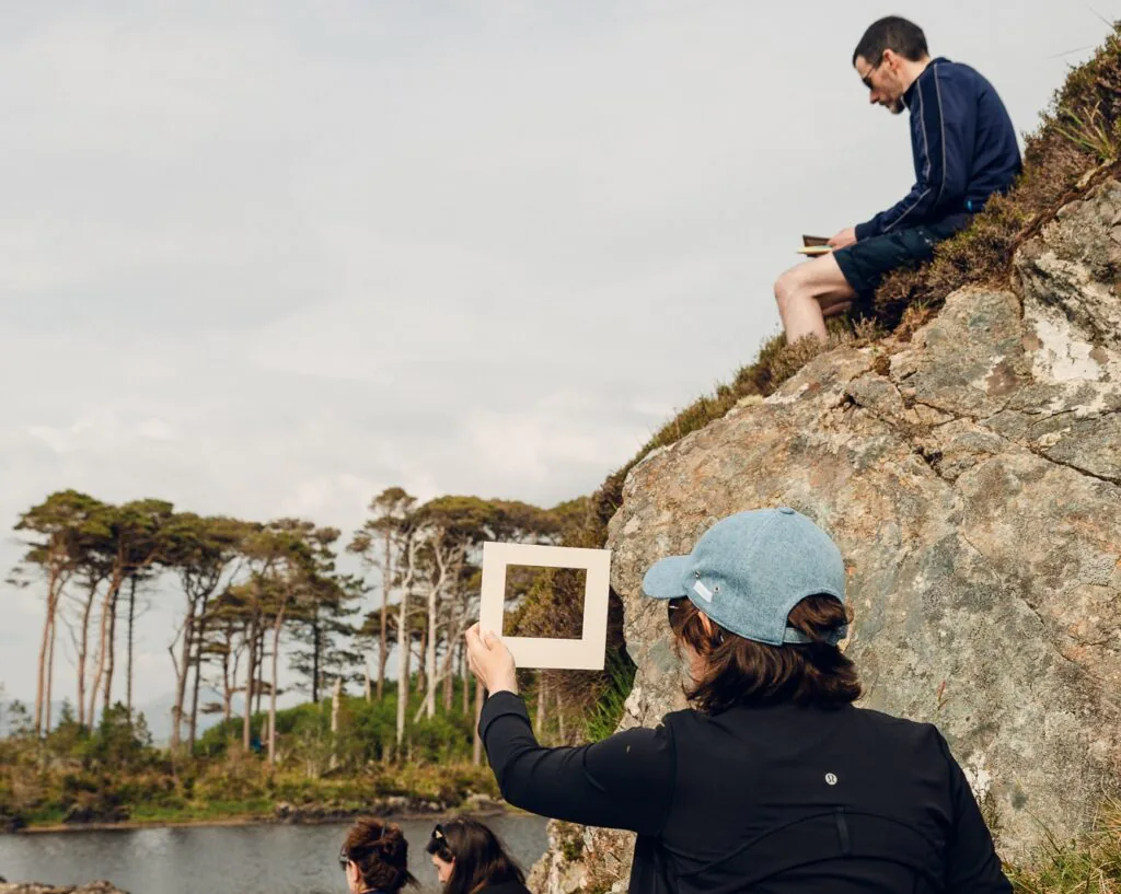 ClareHenderson 050 Several people sit on rocky terrain near water. One person in a blue cap holds up a square frame, partially outlining the landscape. Tall trees stand in the background under a cloudy sky.