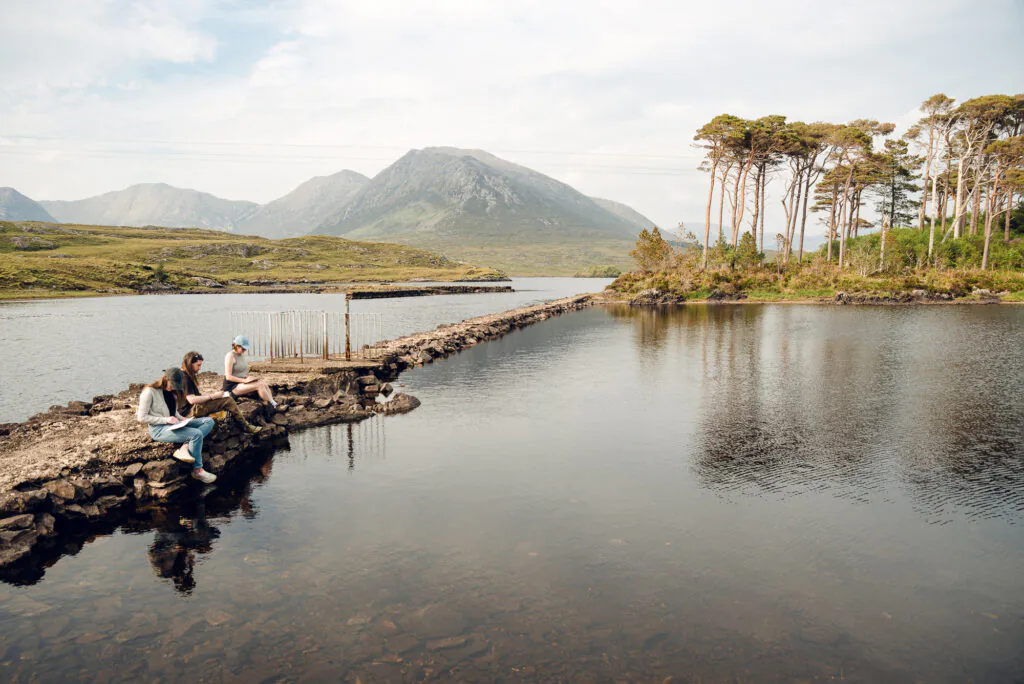 Aoife Herriott Three people sit on a stone path by a calm lake with mountains and trees in the background under a partly cloudy sky.