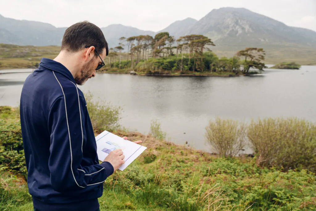 Aoife Herriott A man in a blue jacket sketches on paper while standing by a lakeside with an island and mountains in the background.
