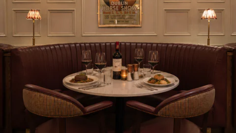 A round restaurant table set for two with wine, water glasses, steaks, and sides, surrounded by a curved burgundy booth, under warm lighting and a Dublin Horse Show poster.