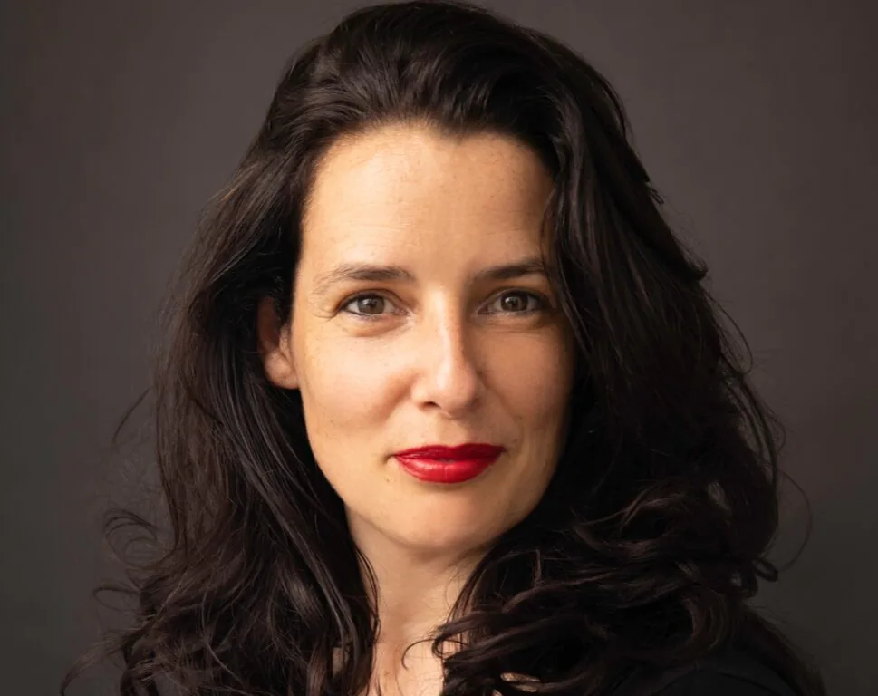 Woman with long dark hair and bright red lipstick poses against a dark background, wearing a black top and looking directly at the camera with a neutral expression.