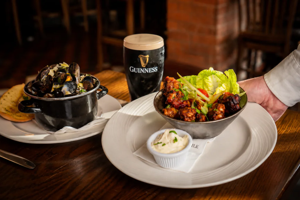 bar mussels and wings A hand serves a bowl of salad and fried food with dip; nearby are a pot of mussels, bread, and a pint of Guinness on a wooden table.