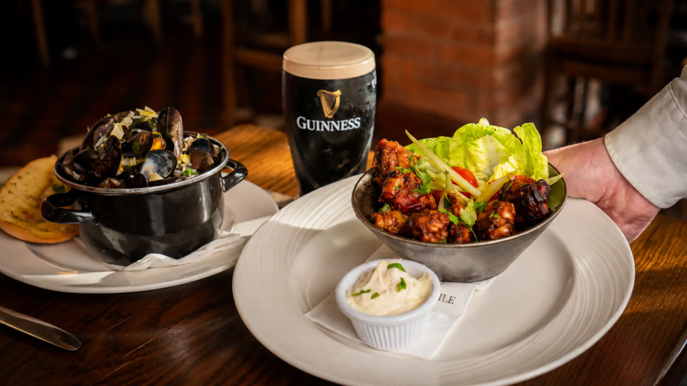 A hand serves a bowl of salad and fried food with dip; nearby are a pot of mussels, bread, and a pint of Guinness on a wooden table.
