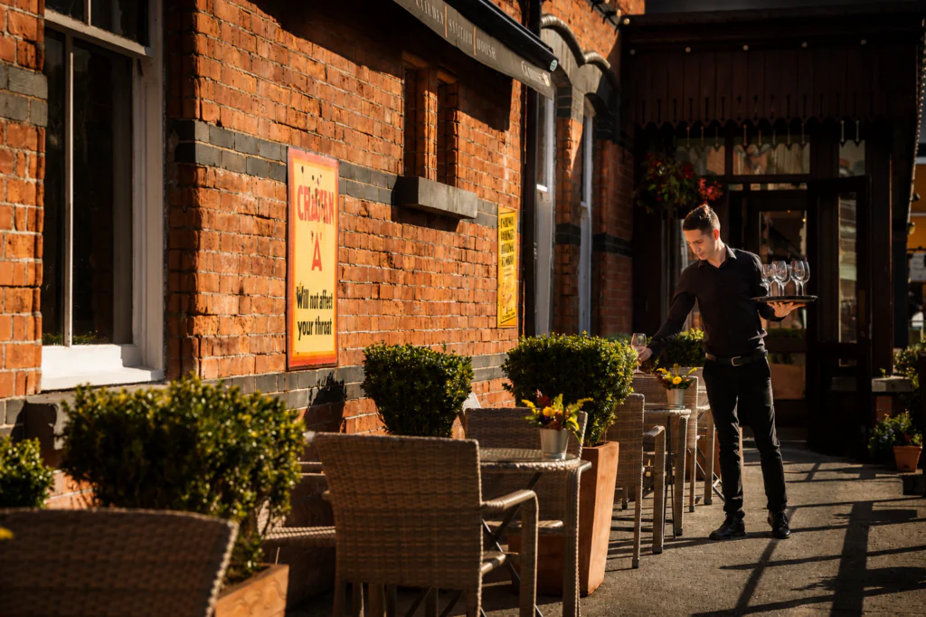 bar outside A waiter in black attire sets a table with glasses on an outdoor patio beside a red brick building lined with wicker chairs and potted plants.