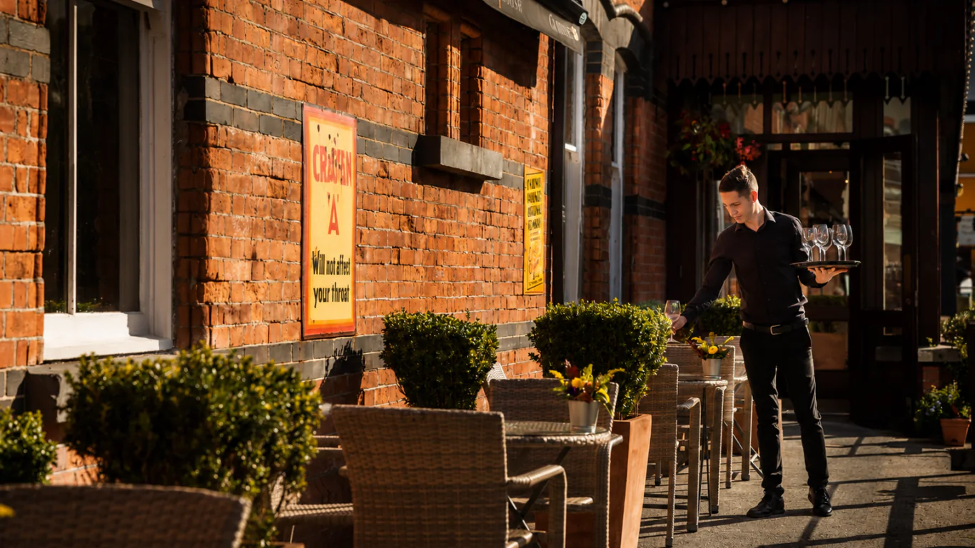 A waiter in black attire sets a table with glasses on an outdoor patio beside a red brick building lined with wicker chairs and potted plants.