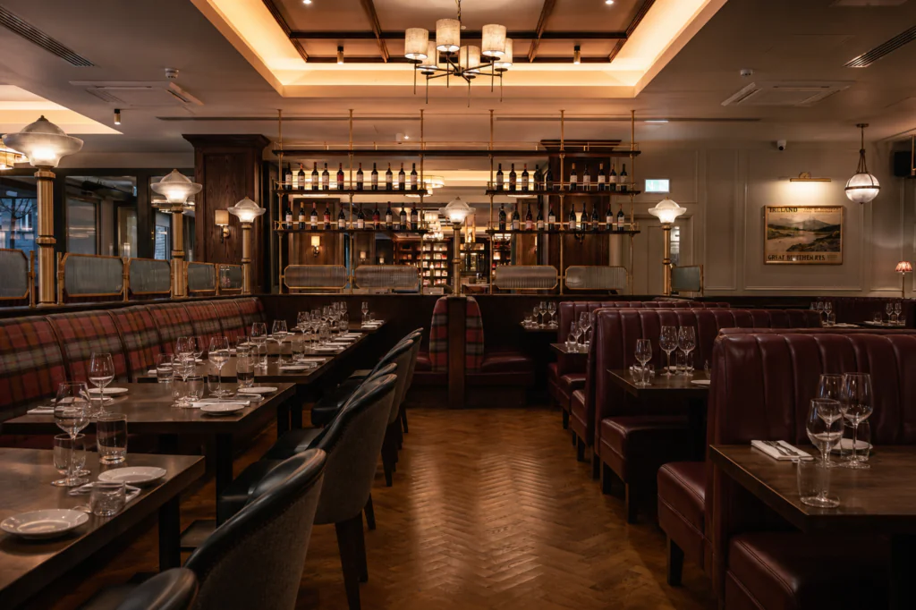 Upscale restaurant interior with empty tables set for dining, leather booths, wooden floors, and a well-lit bar area in the background.