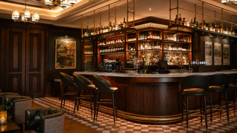 Elegant bar with a curved wooden counter, leather barstools, shelves stocked with bottles, warm lighting, and checkered floor tiles.