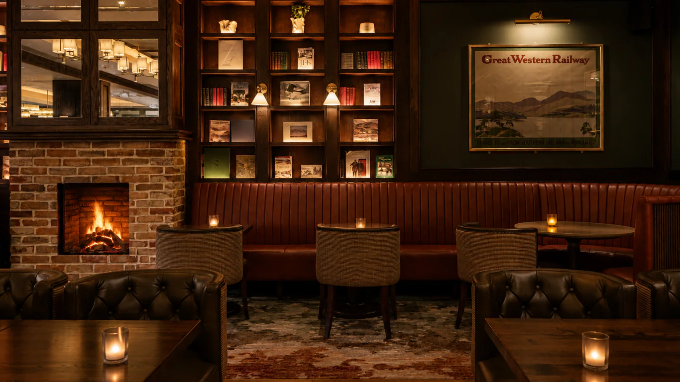 Dimly lit lounge with leather chairs, small tables, a brick fireplace, bookshelves, and a &ldquo;Great Western Railway&rdquo; wall art. Several candles are placed on the tables, creating a cozy atmosphere.
